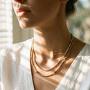 Woman wearing gold layered necklace set at window, white wide-collar top, stone tennis chain and gold ball station chain at collarbone, dappled sunlight on neck

