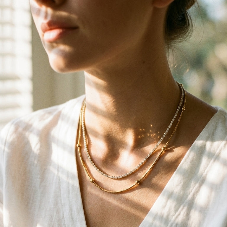 Woman wearing gold layered necklace set at window, white wide-collar top, stone tennis chain and gold ball station chain at collarbone, dappled sunlight on neck
