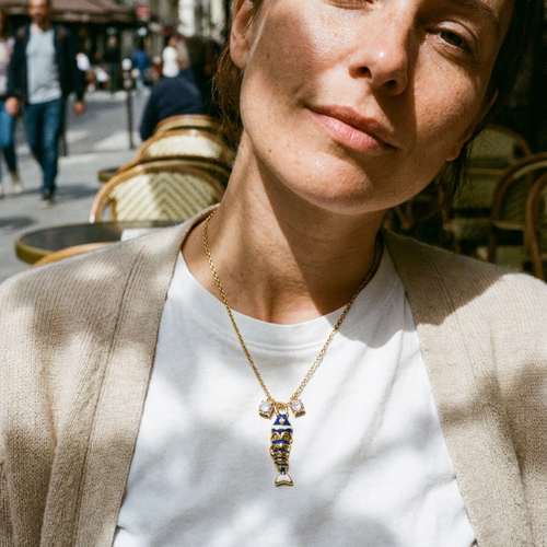 Enamel fish pendant necklace in navy blue worn at outdoor café, gold chain on white tee and beige blazer
