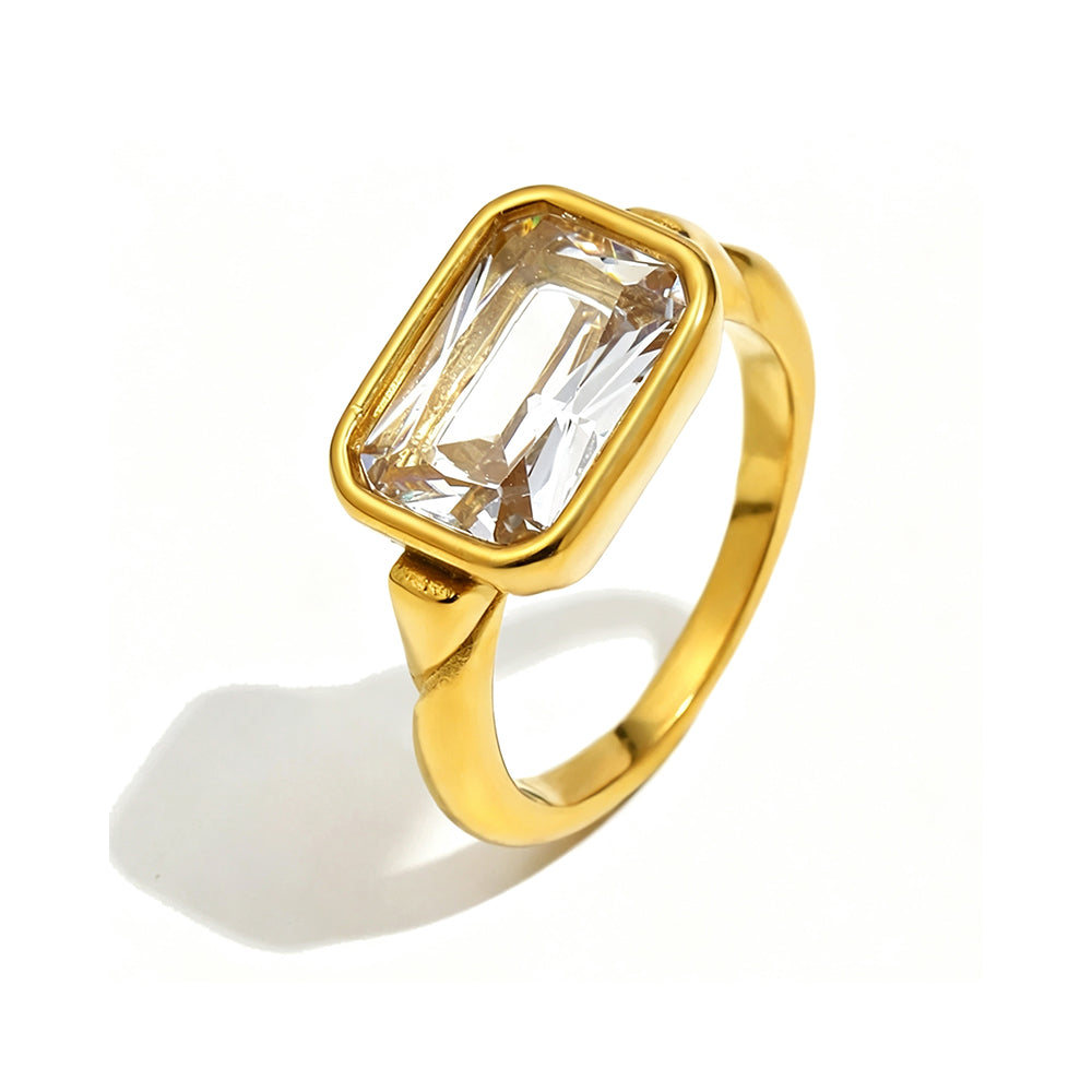 Gold emerald cut stone bezel ring on white background, large clear stone with step-cut facets refracting warm light inside rounded gold bezel, tapered band with shoulder detail, long warm directional shadow
