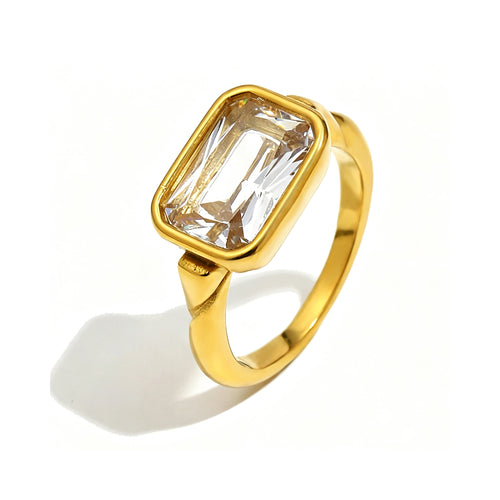Gold emerald cut stone bezel ring on white background, large clear stone with step-cut facets refracting warm light inside rounded gold bezel, tapered band with shoulder detail, long warm directional shadow
