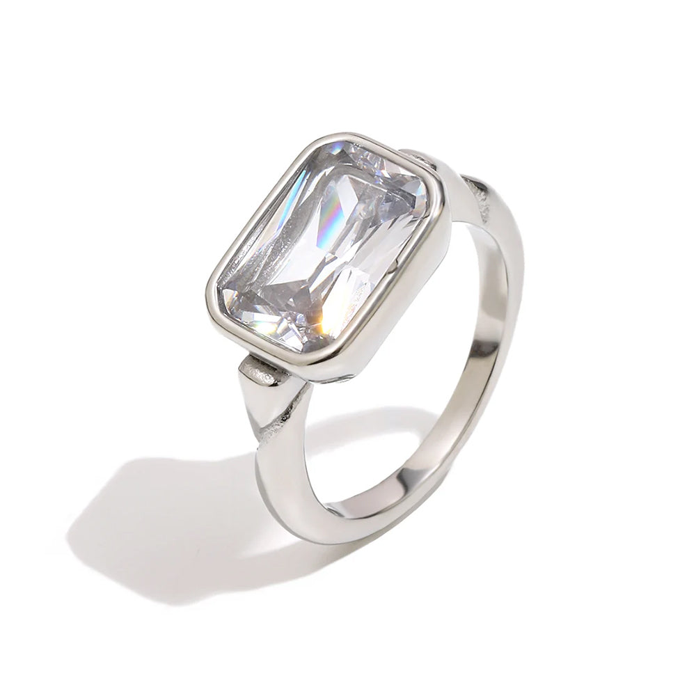 Silver emerald cut stone bezel ring on white background, large clear faceted rectangular stone inside rounded silver bezel, step-cut facets catching cool light, tapered band, soft shadow

