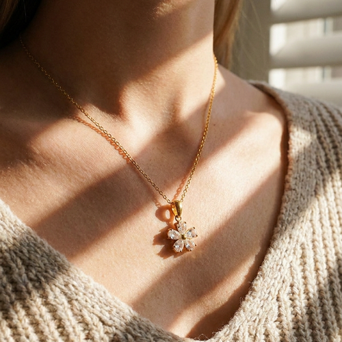 Close-up of gold five petal stone flower necklace at collarbone, five pear-cut clear stones catching warm light through window blinds, beige knit top
