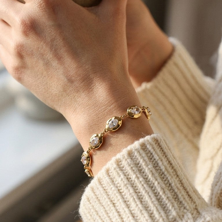 Woman wearing gold hammered link oval stone bracelet, hands clasped together, beige ribbed knit sweater, warm window light with blurred interior background

