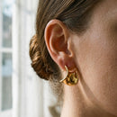Close-up of gold twisted wave stud earring on ear, sculptural folded silhouette and high-polish surface catching soft window light, brown hair in low bun, blurred white window frame in background
