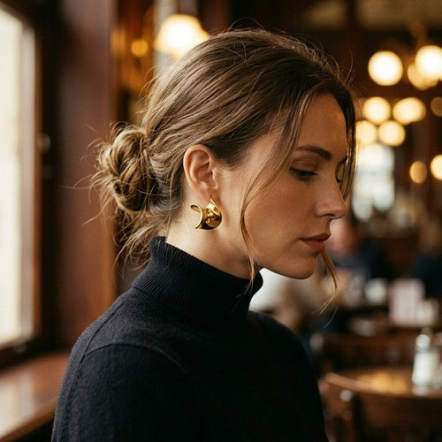 Woman wearing gold twisted wave stud earring at warm-lit cafe, side profile with head slightly down, brown hair in low bun, black turtleneck, blurred golden bokeh lights in background

