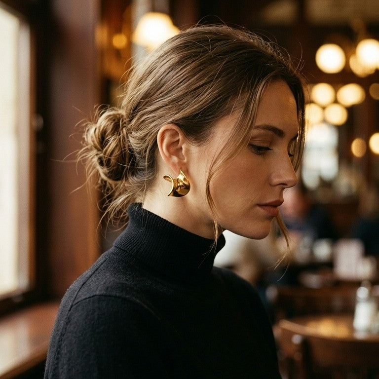 Woman wearing gold twisted wave stud earring at warm-lit cafe, side profile with head slightly down, brown hair in low bun, black turtleneck, blurred golden bokeh lights in background
