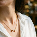 Gold pavé infinity pendant necklace on woman in white shirt, warm indoor light, dainty collarbone necklace
