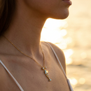 Woman wearing teal enamel fish pendant necklace at beach sunset, gold chain at collarbone, warm golden light
