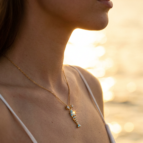 Woman wearing teal enamel fish pendant necklace at beach sunset, gold chain at collarbone, warm golden light
