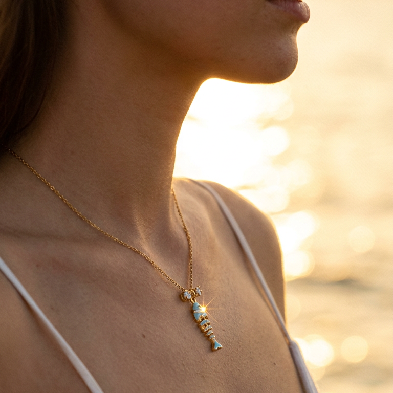 Woman wearing teal enamel fish pendant necklace at beach sunset, gold chain at collarbone, warm golden light
