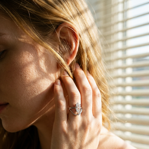 Woman wearing silver marquise crystal double band ring near window, warm morning light through blinds, hand touching hair
