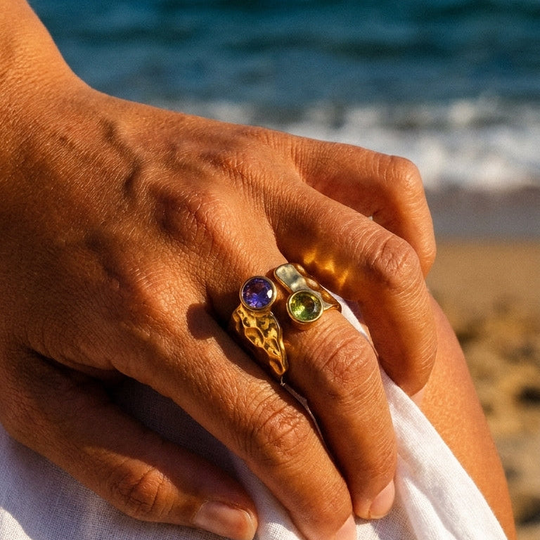 Woman wearing gold dual stone hammered ring at beach sunset, green and purple bezel stones on wide band, warm golden light
