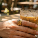 Gold wavy wide band crystal ring worn outdoors, amber center stone catching sunlight, hand holding iced coffee
