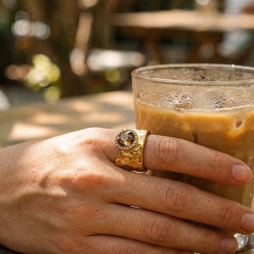 Gold wavy wide band crystal ring worn outdoors, amber center stone catching sunlight, hand holding iced coffee
