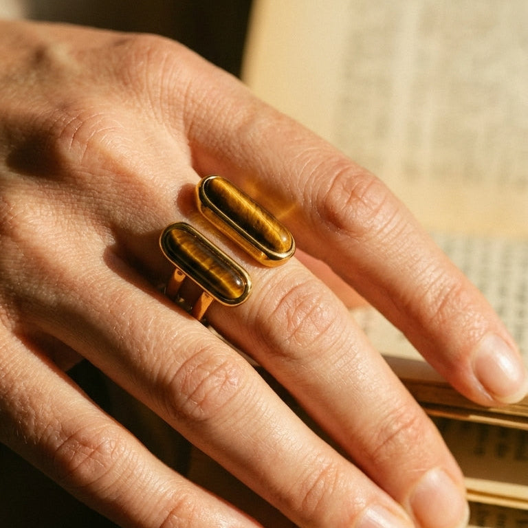 Gold dual tiger's eye open ring worn on index finger resting on vintage book, warm amber stone grain in afternoon light
