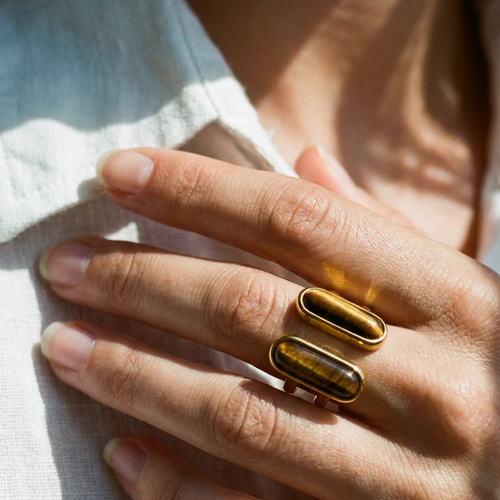 Woman wearing gold dual tiger's eye ring, two warm brown stones on index finger, hands folded over white linen top
