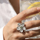 Hand holding iced lemon drink wearing silver hammered texture flower ring, five-petal bloom with green stone center, white linen shirt background
