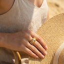 Woman holding straw hat wearing gold hammered oval stone cocktail ring, four champagne stones visible, beige linen top, warm sandy background
