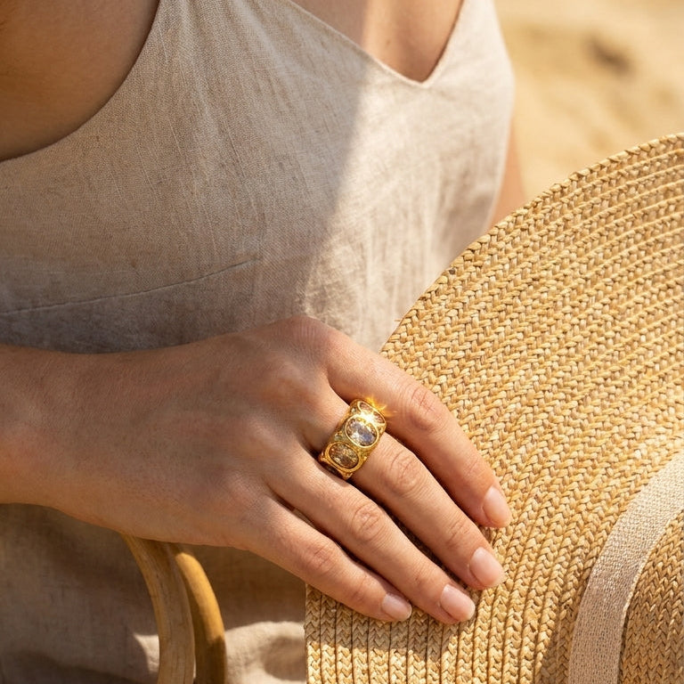 Woman holding straw hat wearing gold hammered oval stone cocktail ring, four champagne stones visible, beige linen top, warm sandy background
