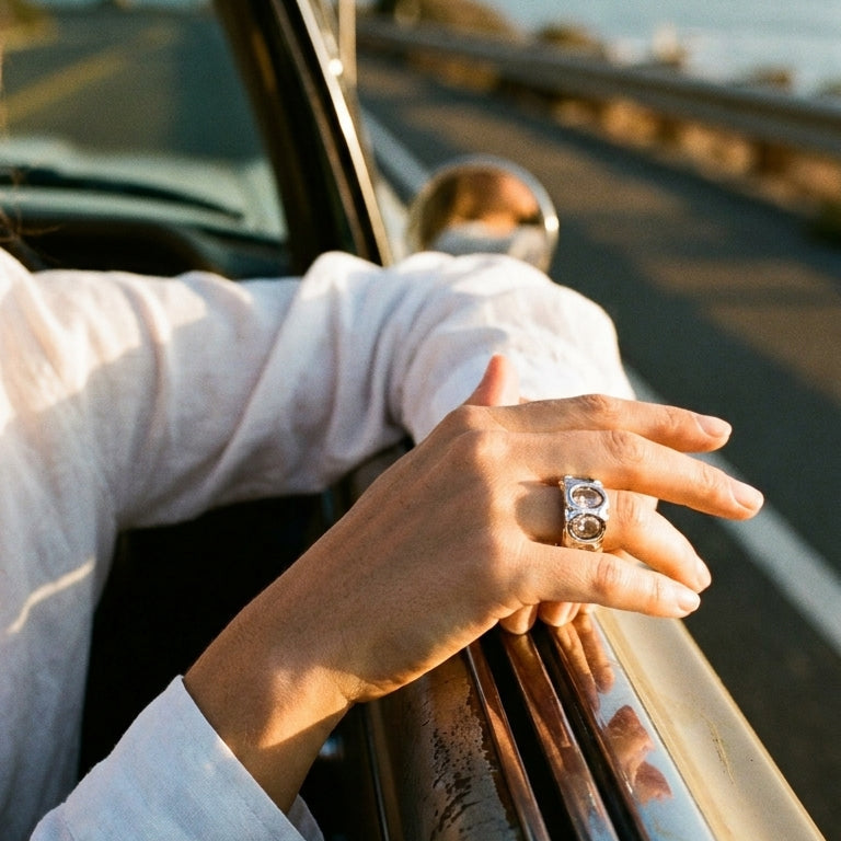 Woman's hand resting on car door wearing silver hammered oval stone cocktail ring, white linen sleeve, golden hour highway background
