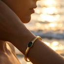 Woman wearing gold oval stone open cuff bracelet in emerald green at sunset beach, wrist raised with cuff catching warm golden light, blurred ocean waves in background
