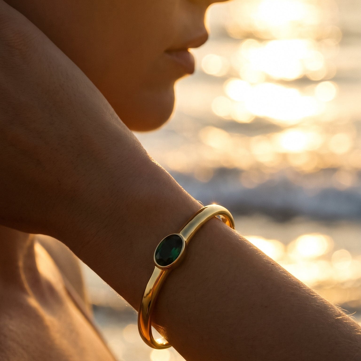 Woman wearing gold oval stone open cuff bracelet in emerald green at sunset beach, wrist raised with cuff catching warm golden light, blurred ocean waves in background
