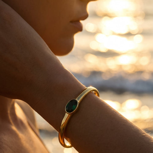 Woman wearing gold oval stone open cuff bracelet in emerald green at sunset beach, wrist raised with cuff catching warm golden light, blurred ocean waves in background

