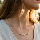 Woman wearing gold pavé wave bar necklace outdoors at golden hour, side profile with loose blonde hair, white linen top, rhinestone wave pendant catching warm backlight, blurred grass in background
