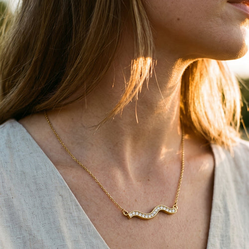 Woman wearing gold pavé wave bar necklace outdoors at golden hour, side profile with loose blonde hair, white linen top, rhinestone wave pendant catching warm backlight, blurred grass in background
