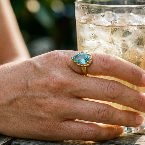 Hand wearing gold blue teardrop stone halo cocktail ring resting on wooden table beside iced drink, aqua blue teardrop stone in multi-prong gold halo setting, warm outdoor light with blurred green foliage background

