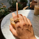 Hand wearing gold marquise stone dome signet ring in brown colorway holding iced coffee glass on marble table, warm amber marquise stone catching direct sunlight, blurred outdoor cafe background
