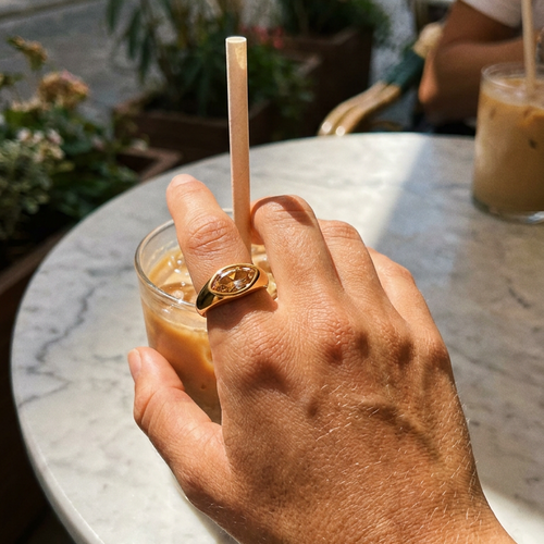 Hand wearing gold marquise stone dome signet ring in brown colorway holding iced coffee glass on marble table, warm amber marquise stone catching direct sunlight, blurred outdoor cafe background

