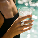 Hand wearing silver oversized emerald cut chunky ring holding glass of iced water, large faceted stone in bright natural light beside water, black tank top, blurred aqua water bokeh background
