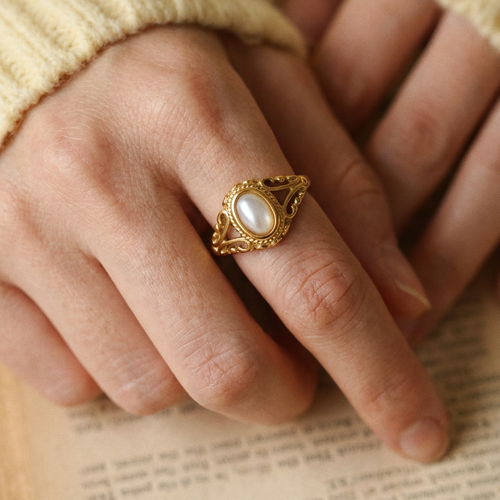Two hands folded over open book pages wearing gold filigree freshwater pearl ring, oval pearl and scrollwork filigree frame in close-up detail, cream knit sweater sleeve, warm indoor light
