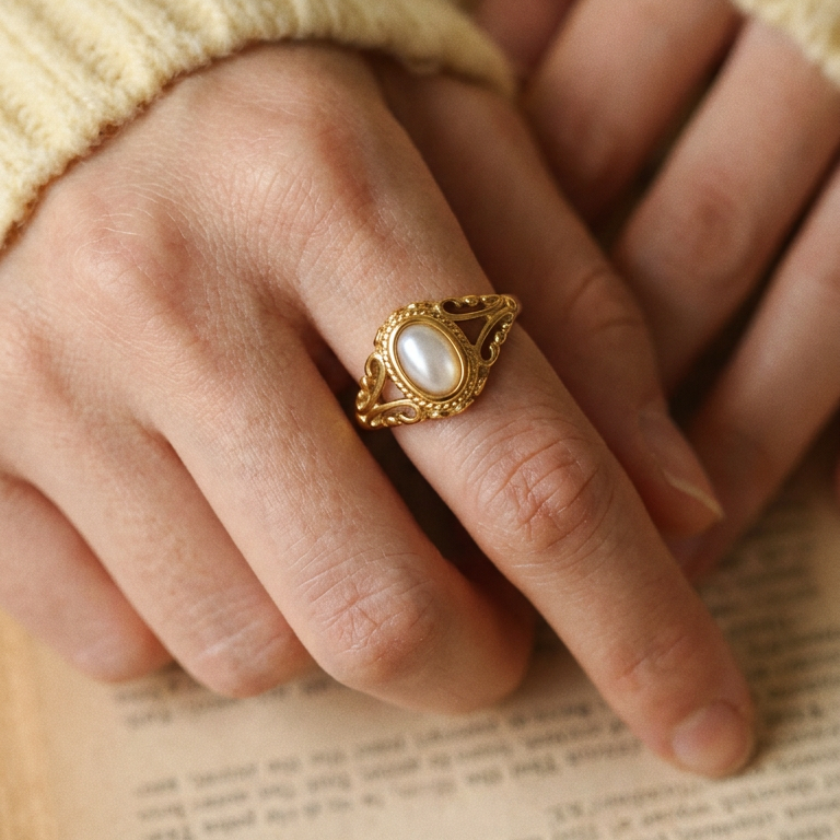 Two hands folded over open book pages wearing gold filigree freshwater pearl ring, oval pearl and scrollwork filigree frame in close-up detail, cream knit sweater sleeve, warm indoor light
