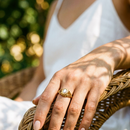 Woman's hand resting on woven rattan chair wearing gold filigree freshwater pearl ring on middle finger, oval pearl and scrollwork setting in dappled garden light, white sundress, blurred green foliage background
