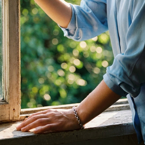 Woman's hand resting on weathered wooden window frame wearing silver textured rolo chain bracelet, hammered round links in cool natural light, light blue linen shirt sleeve rolled up, blurred green foliage background
