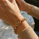 Woman's wrist wearing gold textured rolo chain bracelet, hands clasped together over marble surface, hammered link texture catching warm direct light, cream knit sweater sleeve, blurred background
