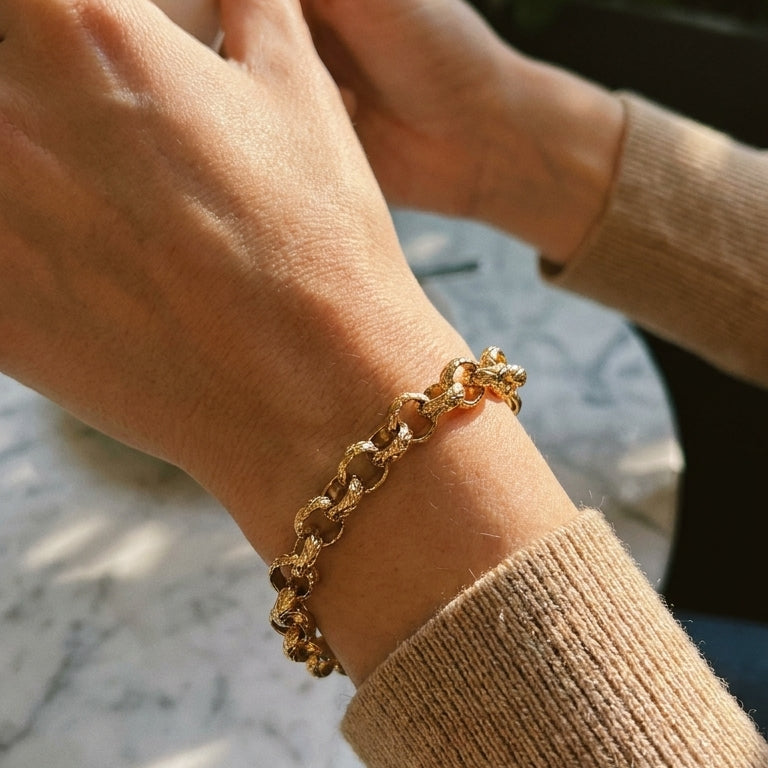 Woman's wrist wearing gold textured rolo chain bracelet, hands clasped together over marble surface, hammered link texture catching warm direct light, cream knit sweater sleeve, blurred background
