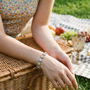 Woman's hands resting on woven picnic basket wearing silver multicolor square stone tennis bracelet, pastel square stones in star-prong settings, floral print top, blurred picnic spread with fruit and glassware in background
