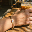 Woman's wrist wearing gold Cuban link bracelet with heart-cut stone holding iced coffee glass on wooden cafe table, heart-shaped stone catching direct sunlight, warm film-grain aesthetic, blurred cafe background
