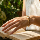Woman's hand resting on open book pages wearing gold Cuban link bracelet with round prong-set stone, white ribbed knit vest, soft natural light, blurred green garden foliage background

