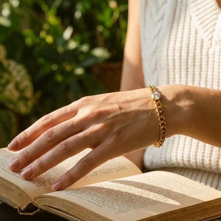 Woman's hand resting on open book pages wearing gold Cuban link bracelet with round prong-set stone, white ribbed knit vest, soft natural light, blurred green garden foliage background
