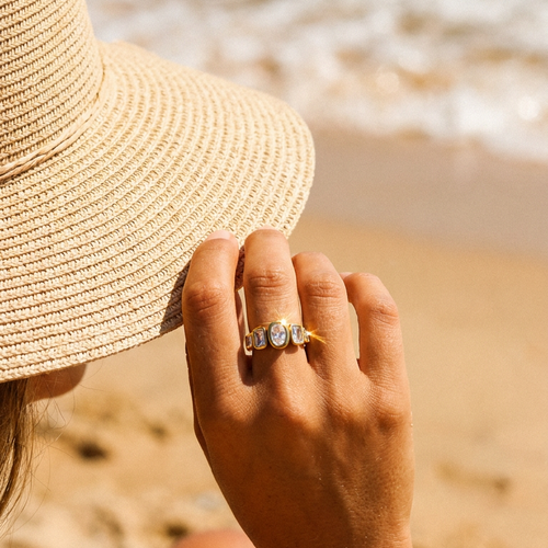 Woman's hand holding straw hat brim on beach wearing gold oval and cushion multi-stone ring on index finger, five bezel-set stones catching bright sunlight, blurred sandy beach and ocean waves background
