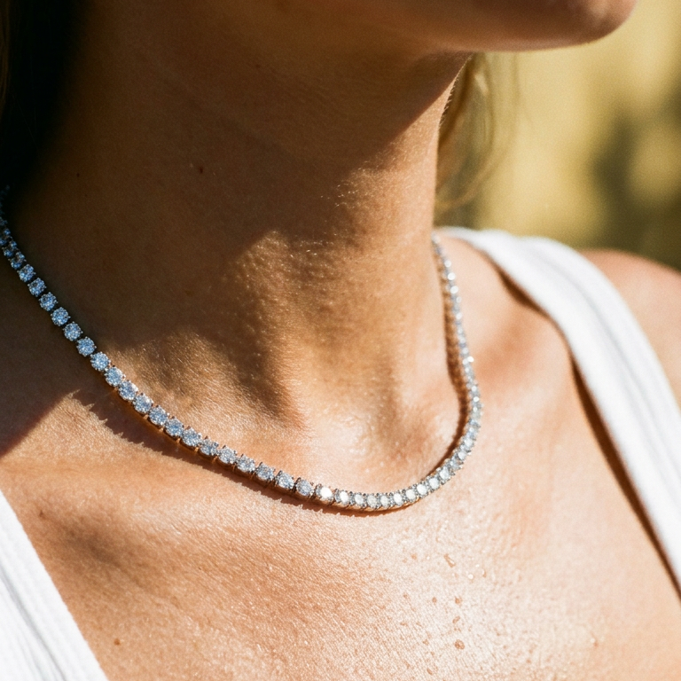 Close-up of woman's collarbone wearing silver round stone tennis necklace, continuous prong-set stones catching warm outdoor sunlight, white tank top neckline, blurred golden foliage background
