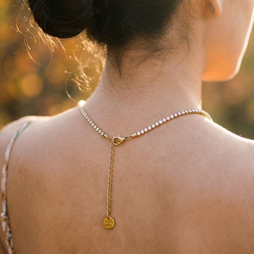 Back view of woman wearing gold round stone tennis necklace, lobster clasp and extender chain with round tag pendant visible at nape of neck, open-back dress, warm sunset bokeh background
