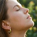 Close-up side profile of woman wearing gold chunky hoop earrings, eyes closed, face tilted upward in warm golden hour sunlight, mirror-polished hoop catching light at earlobe, blurred green foliage background
