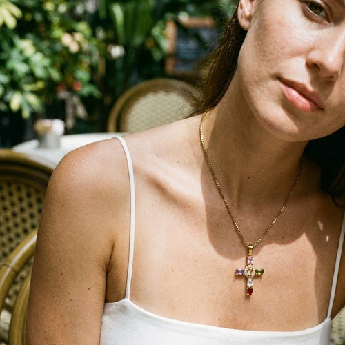 Woman sitting at outdoor cafe wearing gold multicolor stone cross pendant necklace, cross pendant with colored stones visible at collarbone, white spaghetti strap top, blurred rattan chair and tropical foliage background
