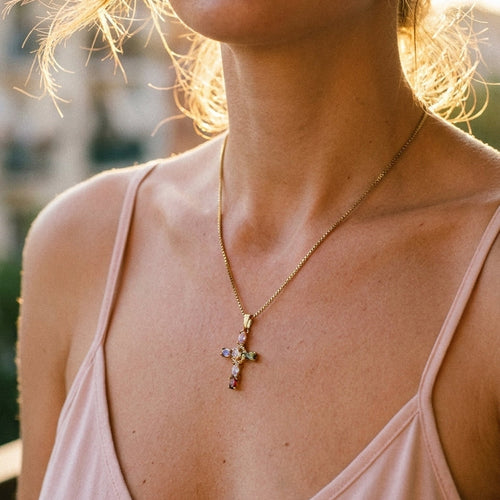 Woman's collarbone wearing gold multicolor stone cross pendant necklace, cross pendant with colored stones catching warm golden hour sunlight, pink slip dress, blurred outdoor background with warm bokeh

